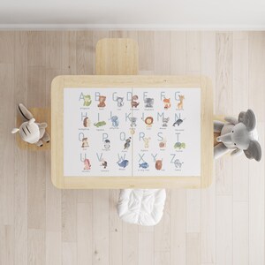 Peut inclure: Un ensemble table et chaise pour enfants en bois clair avec un tableau de l'alphabet illustré d'animaux. Un lapin et un éléphant en peluche sont de chaque côté. Un coussin blanc est sur la chaise.