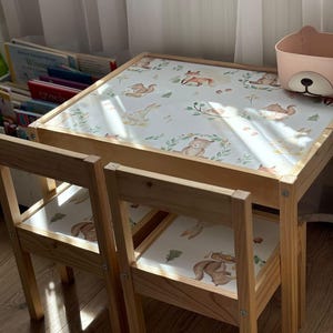 Puede incluir: Una mesa y dos sillas de madera para niños con una mesa blanca con estampado de animales del bosque. La mesa y las sillas están hechas de madera natural y tienen un diseño simple y clásico.