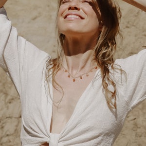 Puede incluir: Una mujer con el pelo largo y rubio lleva una blusa blanca holgada con un nudo en la parte delantera. Lleva un delicado collar de oro con peque&ntilde;os colgantes en forma de l&aacute;grima. La mujer sonr&iacute;e y mira hacia el sol.