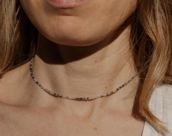 May include: A delicate silver chain necklace with small, multi-colored beads. The beads are a mix of light gray, dark gray, and black.