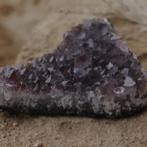 Pode incluir: Um grande aglomerado de cristal de ametista roxo escuro com uma textura áspera e natural. O cristal está sobre uma superfície arenosa.