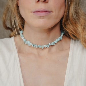 May include: A light blue gemstone choker necklace made of small, irregular shaped stones. The necklace is worn by a person with long, wavy blonde hair.