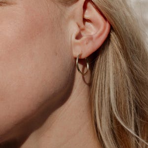 Könnte beinhalten: Nahaufnahme eines Ohrs mit einem goldenen Creolen-Ohrring. Der Ohrring hat ein schlichtes, kreisförmiges Design. Die Person hat helle Haut und blondes Haar. Der Hintergrund ist in einer weichen, neutralen Farbe gehalten.