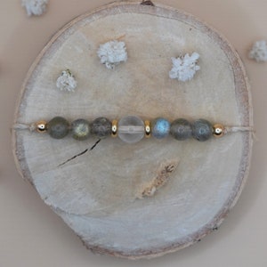 May include: A bracelet made with grey and blue labradorite beads and a clear quartz bead. The bracelet is strung on a thin, natural-colored cord with gold accents.