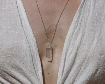 May include: A gold chain necklace with a clear quartz crystal pendant. The necklace is worn by a person wearing a white shirt.