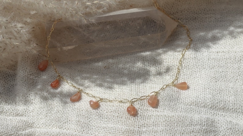 Puede incluir: Un collar de cadena dorada con peque&ntilde;as piedras naranjas en forma de l&aacute;grima que cuelgan. El collar est&aacute; sobre un fondo de tela blanca.