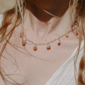 Puede incluir: Un collar de cadena dorada con peque&ntilde;as piedras de cornalina en forma de l&aacute;grima que cuelgan. El collar lo lleva una persona con el pelo largo y rubio ondulado.