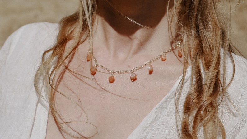 Puede incluir: Un collar de cadena dorada con peque&ntilde;as piedras de cornalina en forma de l&aacute;grima que cuelgan. El collar lo lleva una persona con el pelo largo y rubio ondulado.