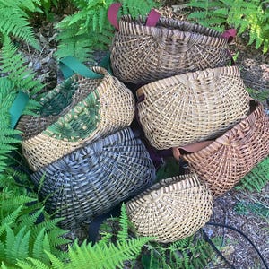 May include: A collection of six woven baskets in various shades of brown, tan, green, and grey. Each basket features a curved, semi-circular shape with a distinct woven pattern and attached fabric straps in colours including red, green, and black.