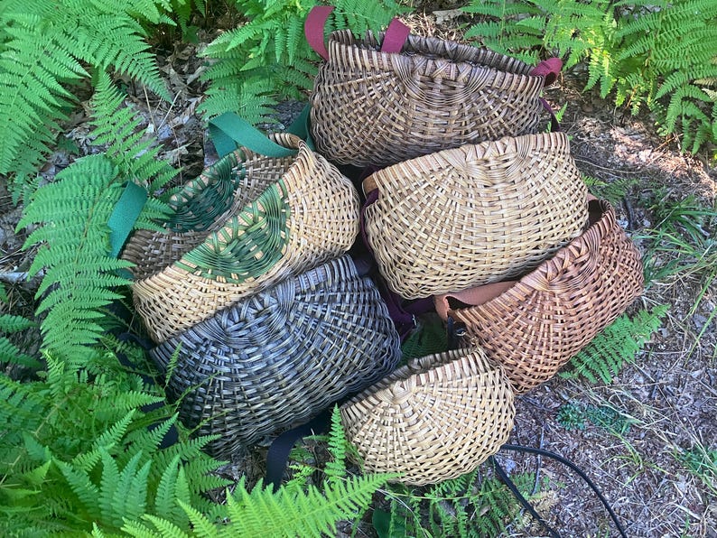 May include: A collection of six woven baskets in various shades of brown, tan, green, and grey. Each basket features a curved, semi-circular shape with a distinct woven pattern and attached fabric straps in colours including red, green, and black.
