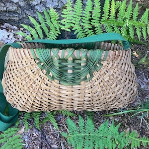 May include: A woven basket bag featuring a natural tan reed body with a decorative green dyed reed accent at the top centre. The basket has a wide, flat green fabric shoulder strap attached to the sides, creating a structured, rounded shape.