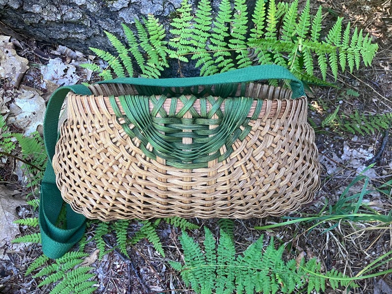 May include: A woven basket bag featuring a natural tan reed body with a decorative green dyed reed accent at the top centre. The basket has a wide, flat green fabric shoulder strap attached to the sides, creating a structured, rounded shape.