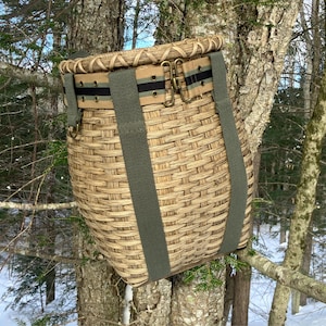 May include: A woven basket with a tan and green color scheme, hanging from a tree branch. The basket has a tan strap with black stripes and green straps for carrying. The basket is made of woven material.