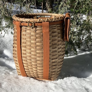 May include: A woven basket with a brown leather strap and buckle closure. The basket has two orange-brown straps that run down the sides. The basket is sitting in the snow with evergreen branches in the background.