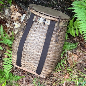May include: A cylindrical woven basket with a flat wooden lid, featuring two vertical black fabric straps, a camouflage-patterned fabric band near the top, a metal buckle closure, and decorative brown wooden beads on thin leather cords.