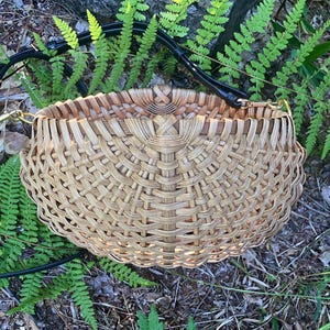 May include: A round, woven basket-style handbag featuring a light tan, natural reed material with a complex, radiating lattice pattern. The bag includes a black leather shoulder strap attached with gold-toned metal hardware clips on each side.