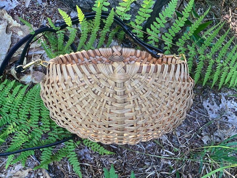 May include: A round, woven basket-style handbag featuring a light tan, natural reed material with a complex, radiating lattice pattern. The bag includes a black leather shoulder strap attached with gold-toned metal hardware clips on each side.