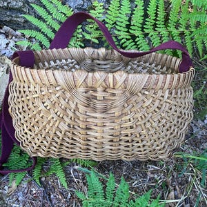 May include: A woven basket with a natural tan finish and a deep burgundy fabric strap. The basket features a structured, rounded rectangular shape with an intricate, interlaced reed pattern throughout the body and a reinforced upper rim.