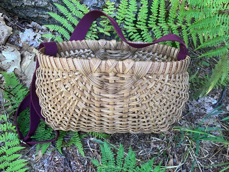 May include: A woven basket with a natural tan finish and a deep burgundy fabric strap. The basket features a structured, rounded rectangular shape with an intricate, interlaced reed pattern throughout the body and a reinforced upper rim.