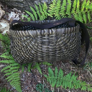 May include: A dark brown woven basket with a rectangular shape and a cross-hatch pattern. The basket features a wide, flat, dark brown fabric strap attached to the sides, allowing it to be carried or worn over the shoulder as a functional bag.