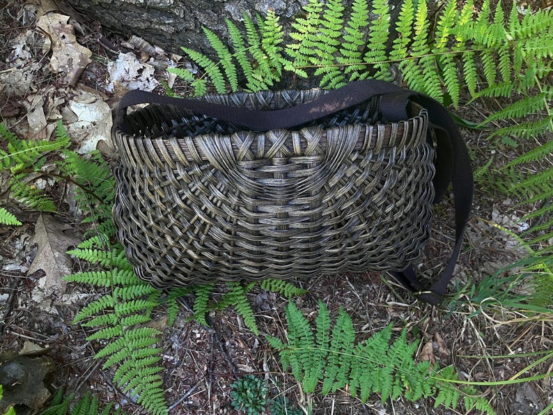 May include: A dark brown woven basket with a rectangular shape and a cross-hatch pattern. The basket features a wide, flat, dark brown fabric strap attached to the sides, allowing it to be carried or worn over the shoulder as a functional bag.