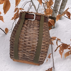 May include: A woven brown basket with a leather belt and silver buckle. The basket has olive green straps and a wooden base. The basket is sitting in the snow with brown leaves and tree branches in the background.