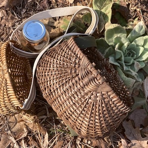 May include: Two woven wicker baskets with a natural brown finish. One basket is round and the other is rectangular with rounded corners. Both baskets feature a light-coloured fabric strap attached to the sides for carrying or hanging purposes.