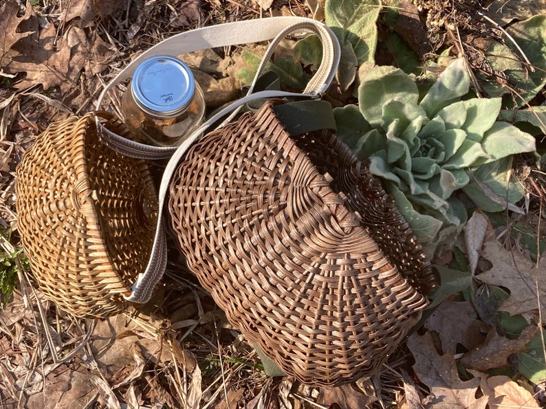 May include: Two woven wicker baskets with a natural brown finish. One basket is round and the other is rectangular with rounded corners. Both baskets feature a light-coloured fabric strap attached to the sides for carrying or hanging purposes.