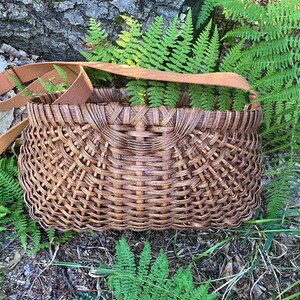May include: A brown woven basket with a curved, open-top design and a wide, flat tan fabric shoulder strap. The basket features a complex, interlocking lattice pattern throughout its body, creating a textured, grid-like appearance in dark brown.