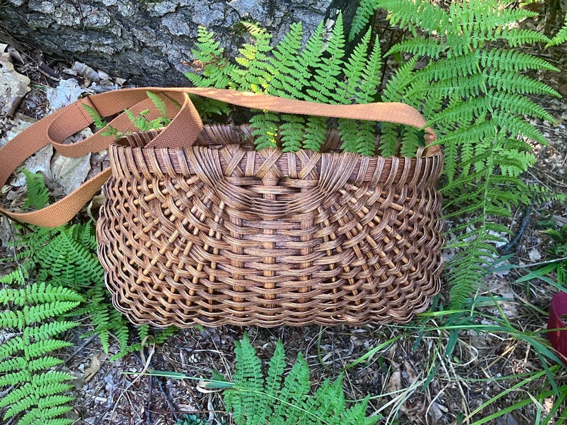 May include: A brown woven basket with a curved, open-top design and a wide, flat tan fabric shoulder strap. The basket features a complex, interlocking lattice pattern throughout its body, creating a textured, grid-like appearance in dark brown.