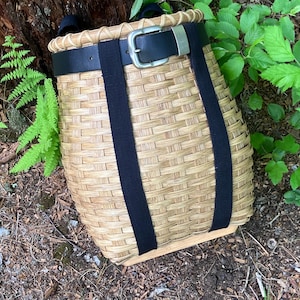 May include: A woven light brown wicker basket with a flat wooden base and a thick black belt strap secured with a silver metal buckle. The basket features a sturdy rim and two vertical black fabric straps extending down the front of the body.