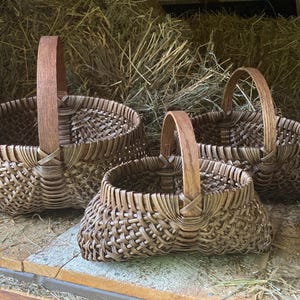 May include: Three dark brown woven baskets with arched wooden handles. Each basket features a complex, interlaced pattern of thin, flexible strips forming a rounded body. The baskets vary in size, arranged in a cluster with visible textures.