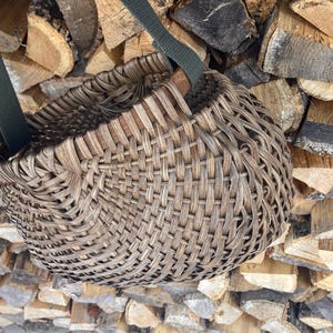 May include: A round, woven basket with a dark brown, weathered finish featuring a complex, interlaced pattern. The basket is equipped with two sturdy, dark olive green fabric straps attached to the rim, allowing for easy carrying or hanging.