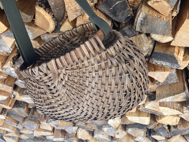 May include: A round, woven basket with a dark brown, weathered finish featuring a complex, interlaced pattern. The basket is equipped with two sturdy, dark olive green fabric straps attached to the rim, allowing for easy carrying or hanging.