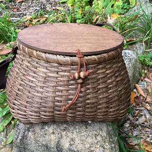 May include: A brown woven basket with a smooth, circular wooden lid. The basket features a dark brown adjustable fabric strap and a decorative leather cord closure with three small wooden beads attached to the front rim.