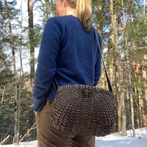 May include: A dark brown woven basket handbag with a rounded, structured shape and a wide, flat top opening. The basket features a tight, intricate lattice weave pattern and is attached to a thin, dark-coloured shoulder strap for carrying.