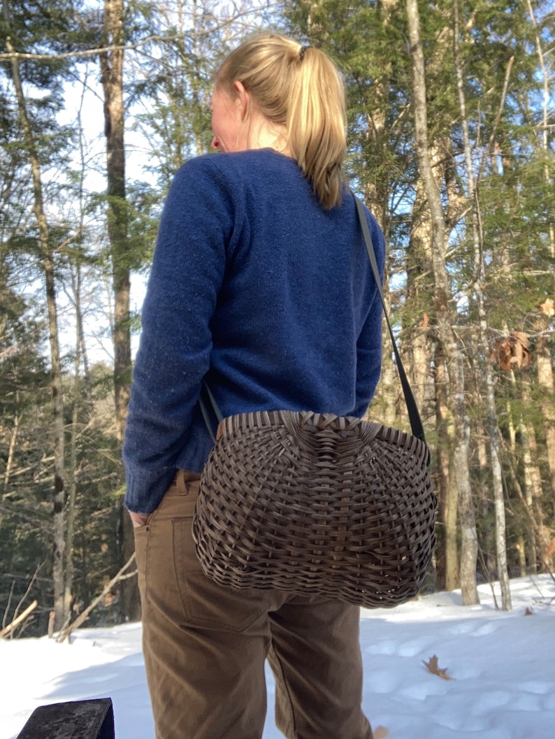 May include: A dark brown woven basket handbag with a rounded, structured shape and a wide, flat top opening. The basket features a tight, intricate lattice weave pattern and is attached to a thin, dark-coloured shoulder strap for carrying.