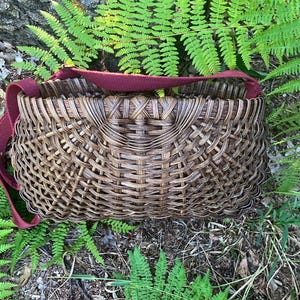 May include: A dark brown woven basket with a complex, interlaced pattern and a wide, flat rim. The basket features a sturdy, solid maroon fabric shoulder strap attached to the sides, allowing for easy carrying of the rounded, open-top container.