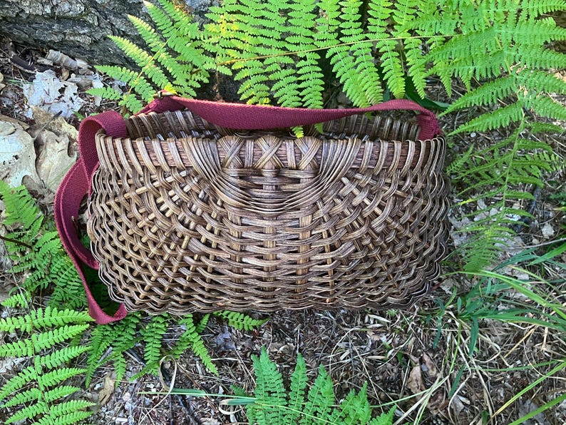 May include: A dark brown woven basket with a complex, interlaced pattern and a wide, flat rim. The basket features a sturdy, solid maroon fabric shoulder strap attached to the sides, allowing for easy carrying of the rounded, open-top container.