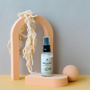May include: A clear glass spray bottle with a black cap and a label that reads "Desert Flora Linen and Body Spray" sits on a peach-colored platform. The bottle is positioned in front of a peach-colored arch and a beige sphere.