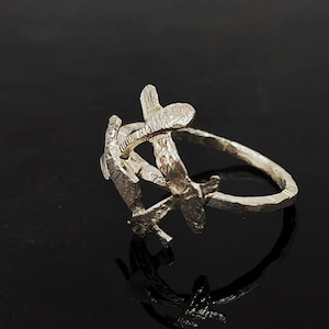 Puede incluir: Un anillo de plata con un diseño texturizado orgánico que se asemeja a ramas o ramitas. El anillo está abierto y tiene un acabado martillado.