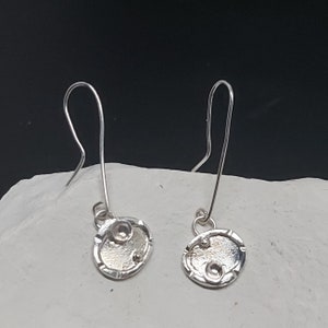 Puede incluir: Un par de pendientes de plata con forma ovalada texturizada y tres pequeños círculos de plata.