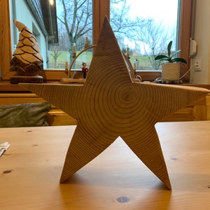 May include: A large, wooden, five-pointed star with a natural wood finish. The star is made from a single piece of wood and has visible growth rings.