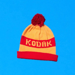 May include: A yellow and red knitted beanie with a red pom pom on top. The beanie has the word "KODAK" written in yellow across the front.