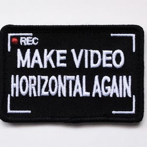 Op de afbeelding: Zwarte geborduurde patch met witte tekst die "MAKE VIDEO HORIZONTAL AGAIN" luidt, binnen een wit rechthoek met een rode cirkel die "REC" in de linkerbovenhoek heeft.