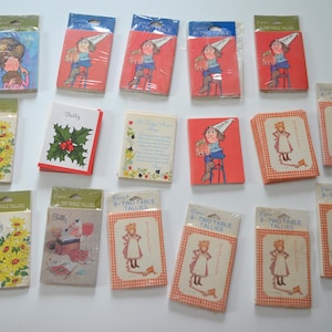 May include: A collection of 16 vintage table tally cards featuring various illustrations. The cards are printed on white cardstock with colorful designs. Some cards feature a holly sprig, a girl in a dress, and a girl in a hat. The cards are labeled "Tally" and "8 Two Table Tallies".