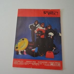 以下が含まれることがあります： 赤と青のデザインのヴィンテージ雑誌の表紙。表紙には、冬のギア、スノーチューブ、荷物を持った3人の人がいます。「SHADE TREE」というテキストが上部にあります。