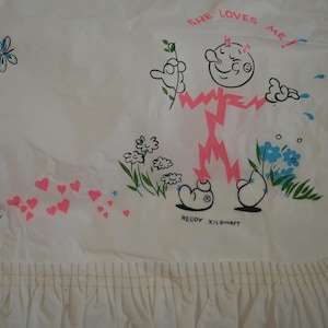 May include: A white fabric item with a ruffled edge featuring a graphic of the character Reddy Kilowatt in pink and black. The design includes the text SHE LOVES ME, a blue butterfly, pink hearts, green stems, and blue flowers.