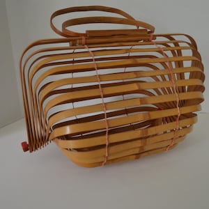 Puede incluir: Un bolso de mano de bambú marrón claro con un diseño único en forma de jaula. El bolso tiene un asa y está sujeto con una fina cuerda de color claro. Tiene un cierre rojo en el lateral.