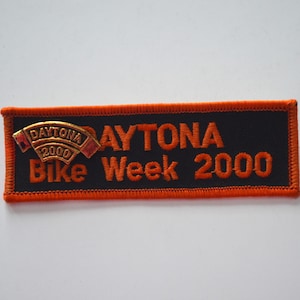 Puede incluir: Parche rectangular negro con bordado y texto en naranja. El texto "DAYTONA Bike Week 2000" está bordado en naranja. Un pin dorado con "DAYTONA 2000" está adjunto. Un artículo de recuerdo.